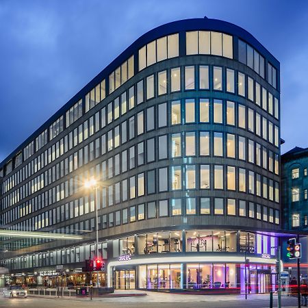 Yotel Глазго Екстер'єр фото The photo shows a modern office building with a distinctive curved shape and a facade made of large glass windows. It is illuminated against a twilight sky, giving it a contemporary appearance. The ground floor features large glass entrance areas, li